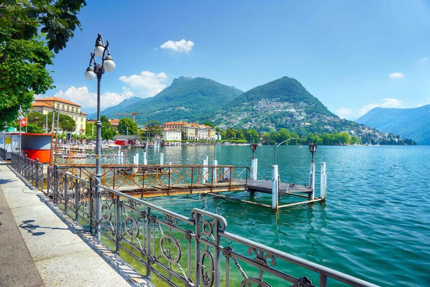 A lakeside promenade with a metal railing, a dock, and buildings in the background. 2