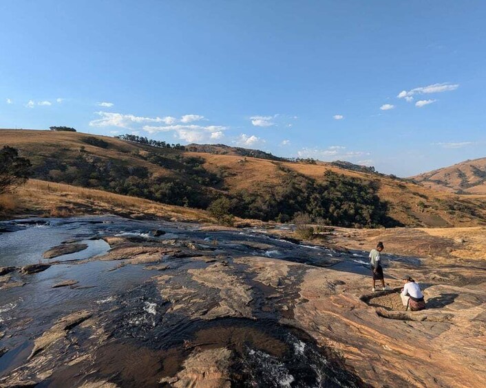 A rocky riverbed with a person sitting and another standing, surrounded by dry hills and sparse vegetation. 5