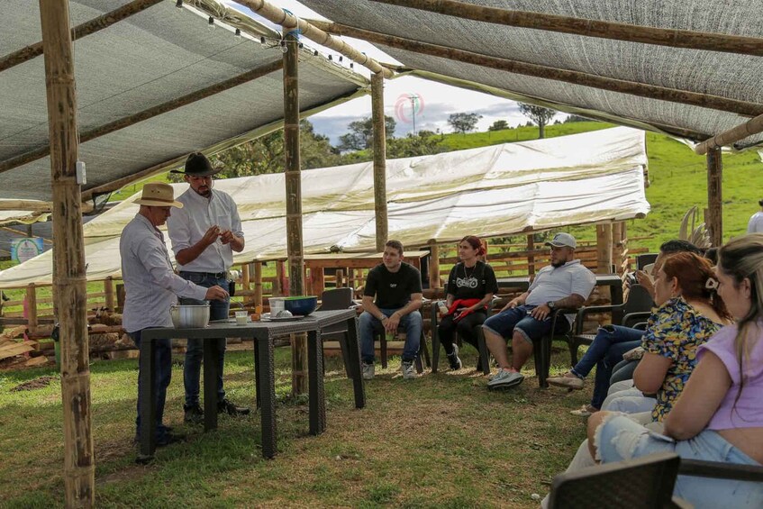 A group of people seated under a shaded area with a table where two individuals are preparing food. 3