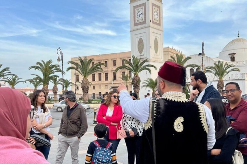 A group of people gathered in a public square with a clock tower and palm trees in the background. 1