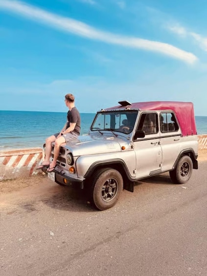 A silver jeep with a red cover parked by the roadside near a body of water. 3