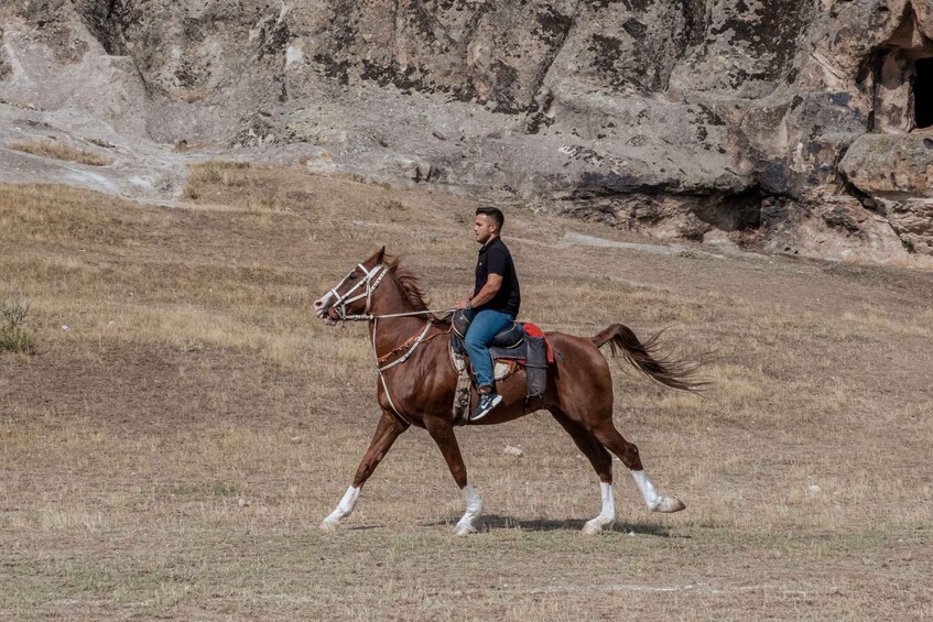 A person riding a horse on a grassy field with rocky terrain in the background. 1