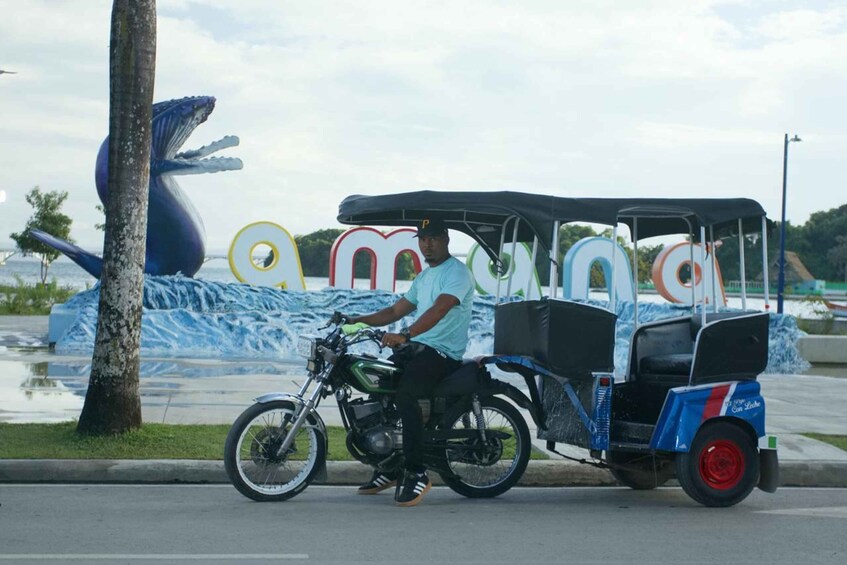 A man on a motorcycle with a tricycle trailer, a large whale sculpture, and a mural with colorful letters. 1