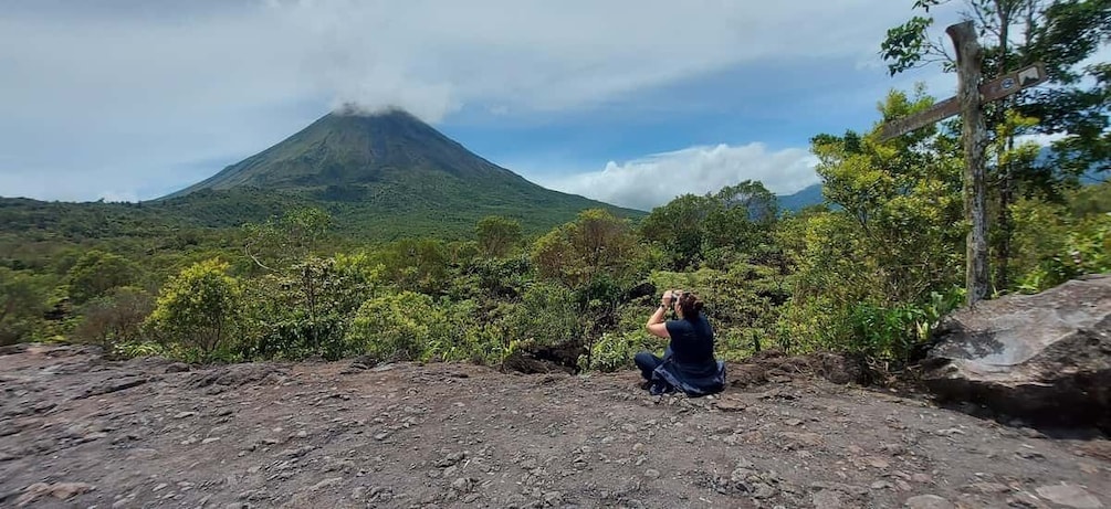 A person taking a photo of a volcano with a forested landscape in the foreground. 3