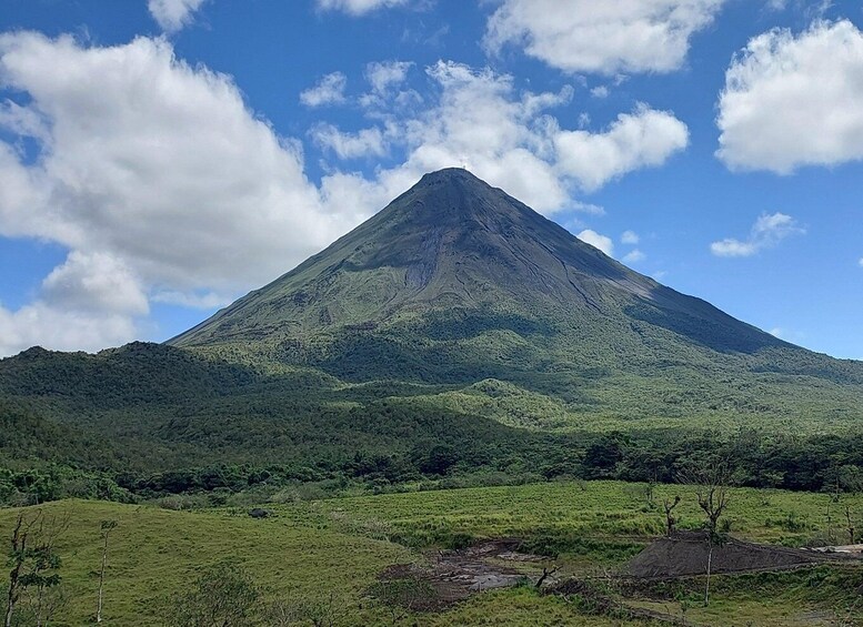 A mountain with a conical peak, surrounded by lush green vegetation and a clear blue sky with scattered clouds. 1