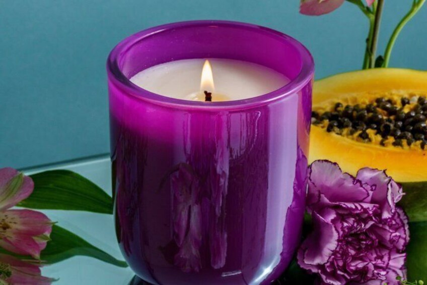 A purple candle in a glass holder with a lit flame, surrounded by flowers and a half-cut papaya. 1