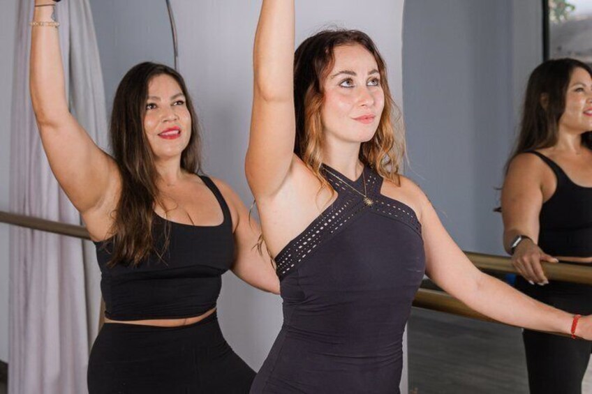 Two women in black workout attire performing a stretching exercise at a barre. 3