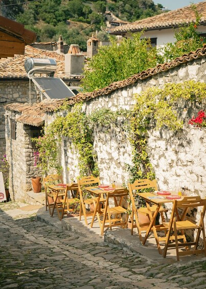 A cobblestone street with wooden tables and chairs, a stone wall with greenery, and traditional buildings in the background. 2