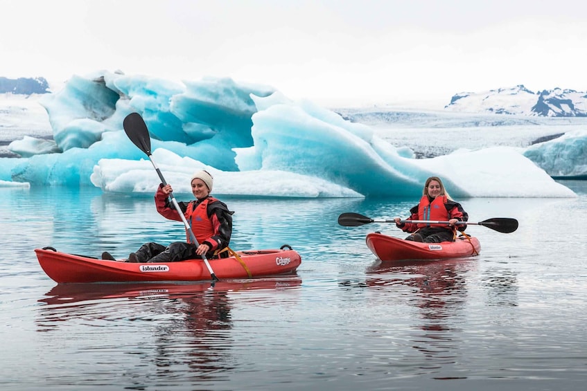 Two people kayaking on calm water with large icebergs in the background. 2