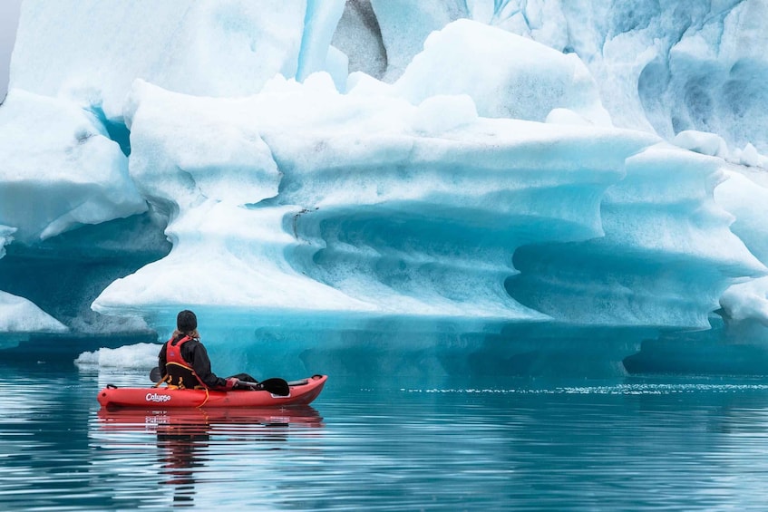 A person in a red kayak on calm water with large icebergs in the background. 1
