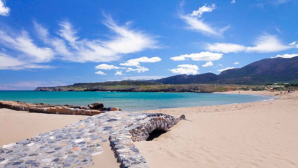 A coastal beach with a stone wall, clear turquoise water, and distant mountains under a blue sky. 5