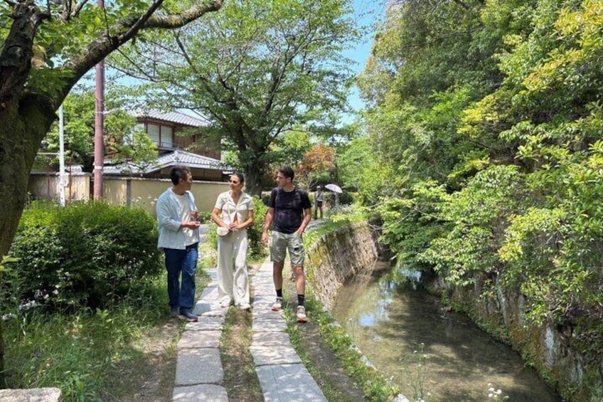 A narrow waterway flanked by lush greenery and stone walls, with people walking along the path. 1
