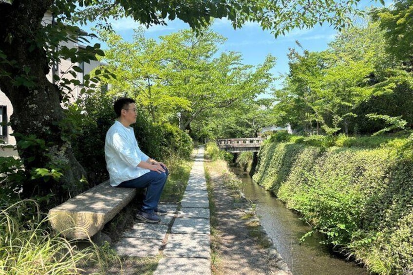 A person sitting on a bench by a canal, surrounded by greenery and a bridge in the background. 5