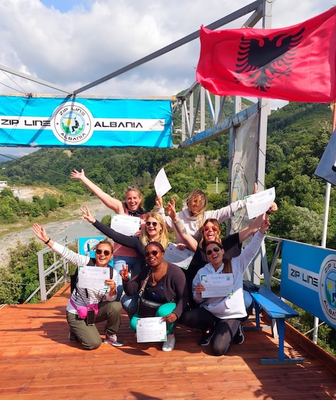 A group of people on a wooden platform with a scenic view of a river and hills in the background, holding certificates and a red Albanian flag. 4