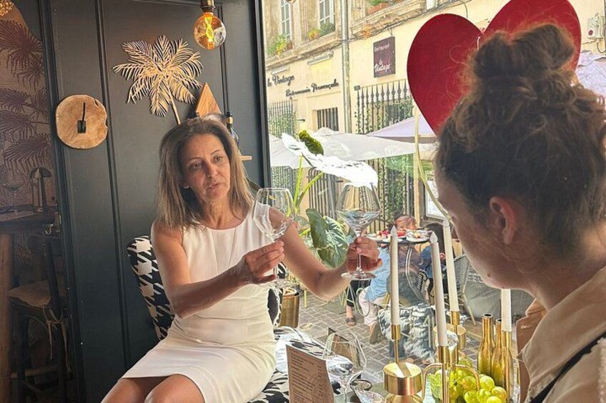 A woman in a white dress sitting at an outdoor café with a table set with wine glasses, bottles, and a fruit basket. 3
