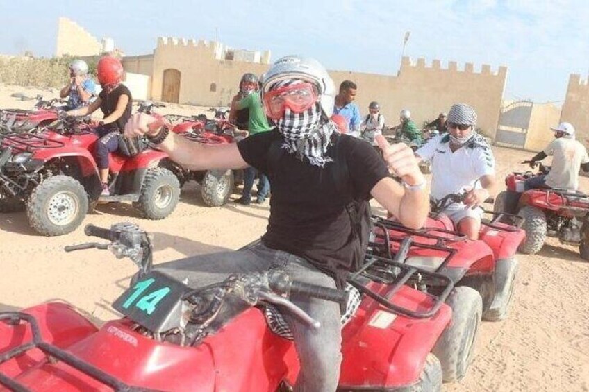 A group of people on red ATVs in a desert-like environment with sandy terrain and a building with a tower in the background. 2