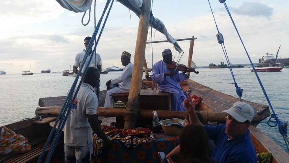 A group of people on a boat with a wooden deck, ropes, and a person playing a stringed instrument. 3