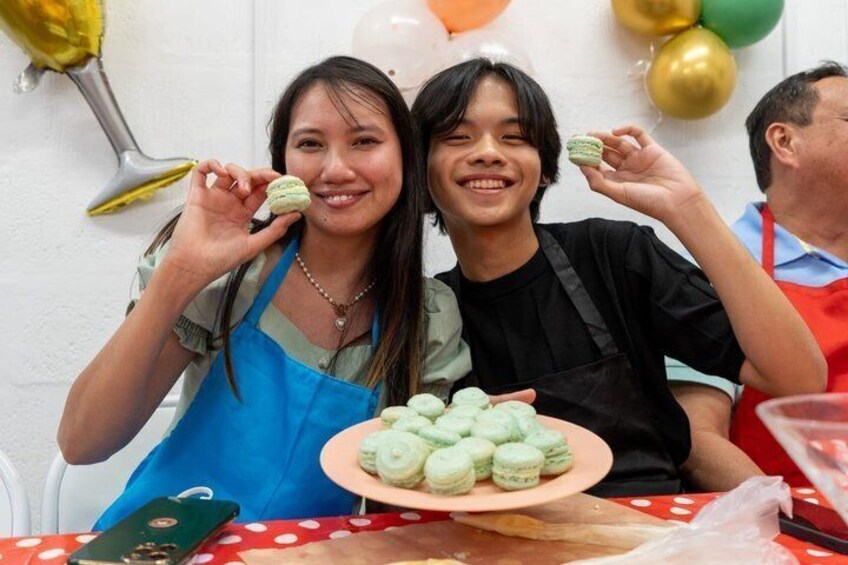 Two people holding macarons at a table with a plate of macarons. 1