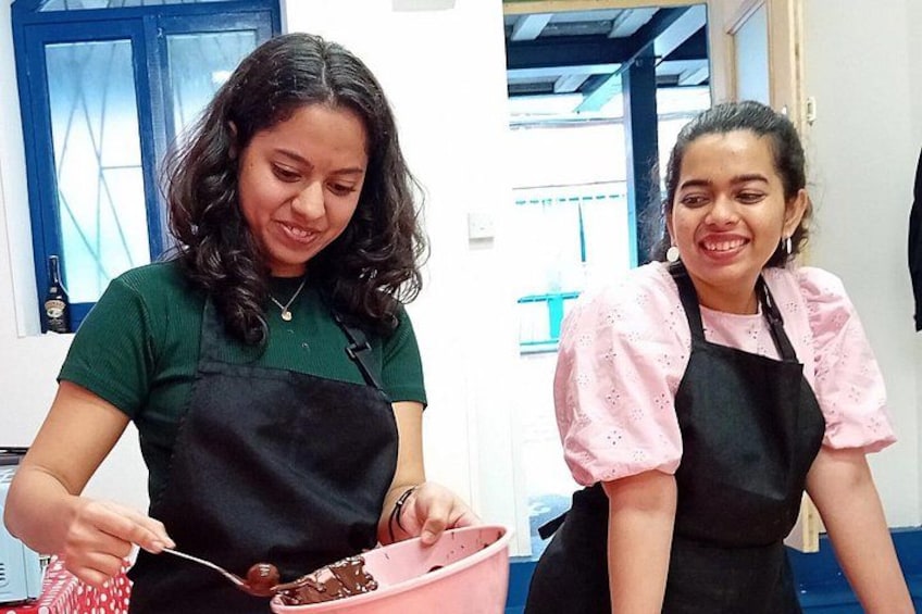 Two individuals wearing aprons, one in a green shirt and the other in a pink shirt, engaged in an activity with a pink bowl and a spoon. 1
