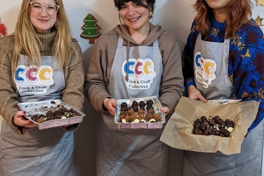 Three people wearing aprons with the 'Cook & Craft Collective' logo, holding trays of food items. 5