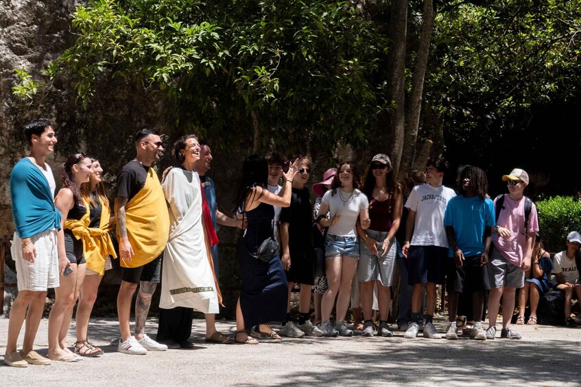 A group of people standing outdoors, some wearing casual clothing, with trees and a rocky background. 2