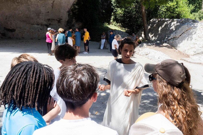 A group of people gathered around a person in a white robe, possibly a guide, in a rocky outdoor setting with trees. 3