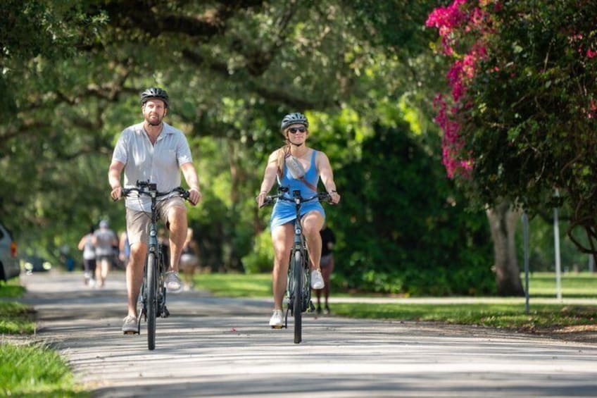 Two cyclists on a paved path in a park with trees and a pink flowering bush. 1