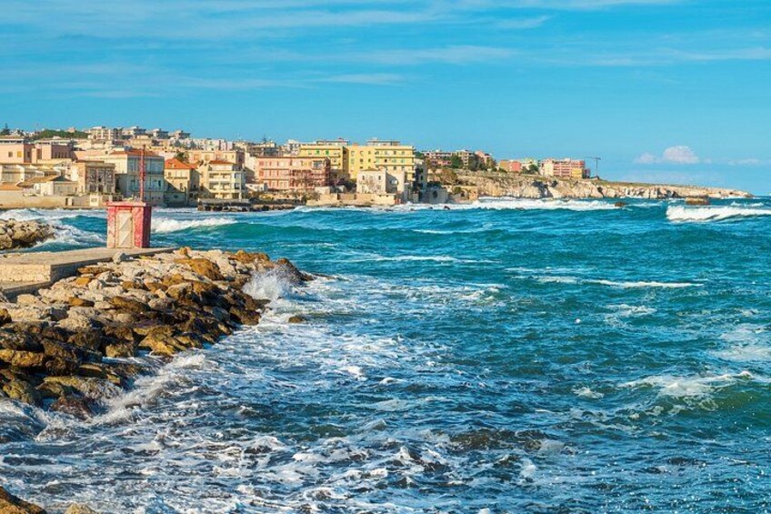 A coastal town with buildings along the shore, waves crashing onto rocks, and a clear blue sky. 2