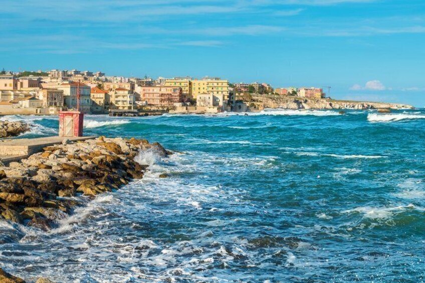 A coastal town with buildings along the shore, waves crashing onto the rocks, and a clear blue sky. 3