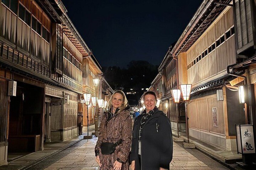 A narrow street with wooden buildings, street lamps, and a person standing in the middle. 3