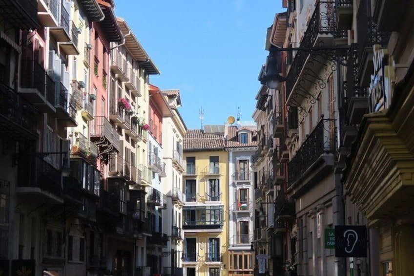 A narrow street flanked by multi-story buildings with balconies, a clear blue sky, and a visible sign indicating a tax office. 2