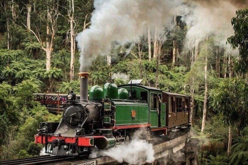 A steam locomotive with green and red carriages traveling through a forested area. 1