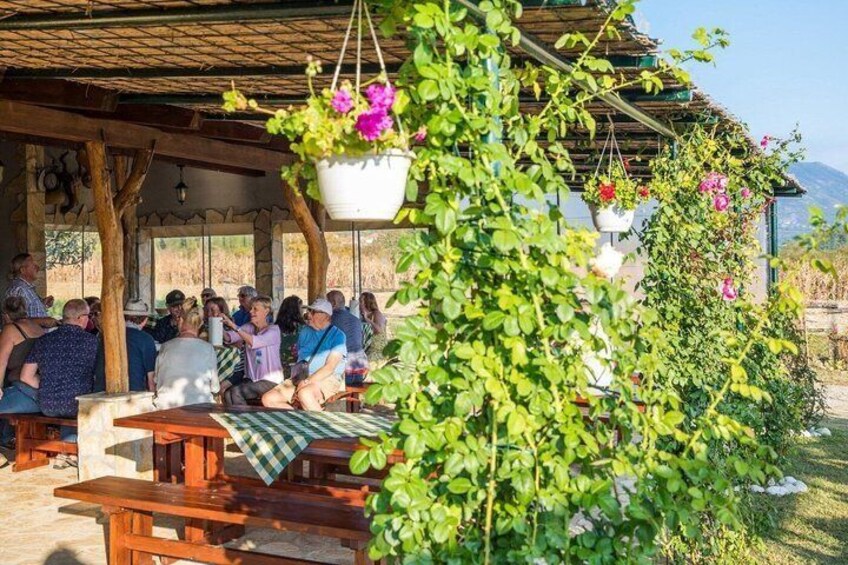 A group of people seated at wooden tables under a covered area with hanging flower baskets. 1