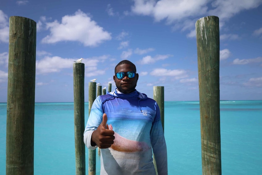 A person giving a thumbs-up gesture on a wooden pier with clear turquoise water in the background. 2