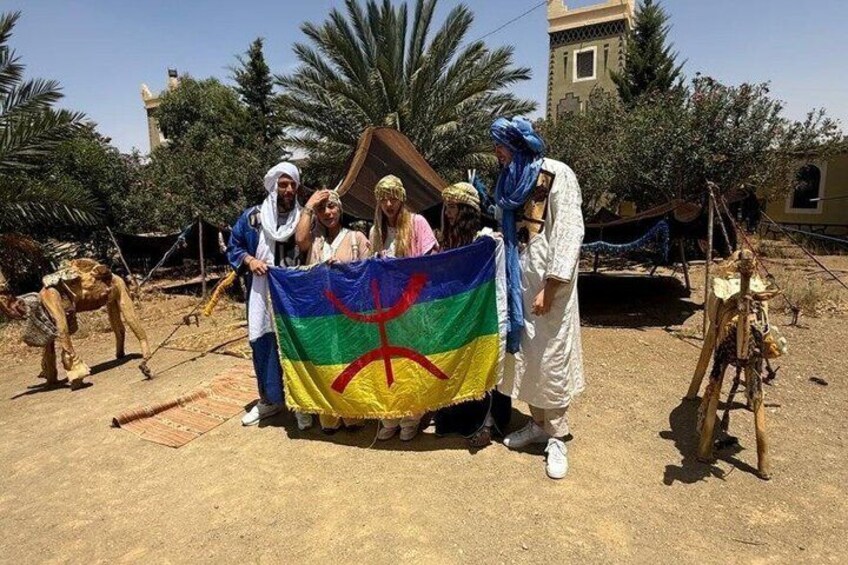 A group of people holding a flag with a peace symbol, standing in a desert-like area with palm trees and a building in the background. 5
