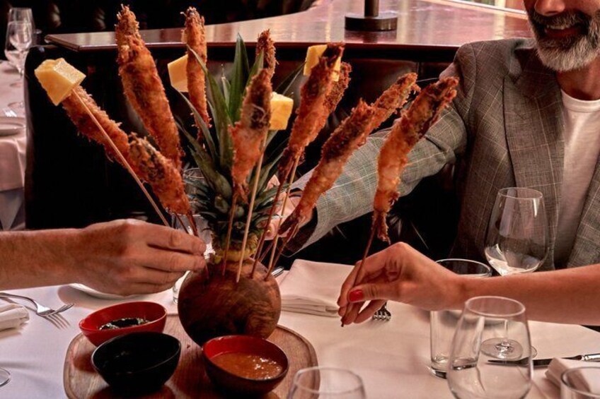 A dining table with a centerpiece of skewered food, a pineapple, and a bowl of sauce. 4