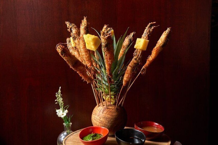 A wooden vase with fried plantains, a small vase with white flowers, and two bowls with sauces on a wooden table. 3