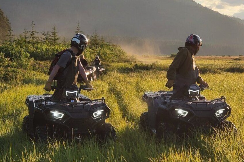 Two people on ATVs in a grassy field with a forest in the background. 1