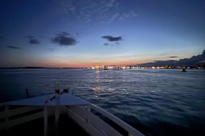 A calm sea at dusk with a boat docked in the foreground and city lights in the distance. 2