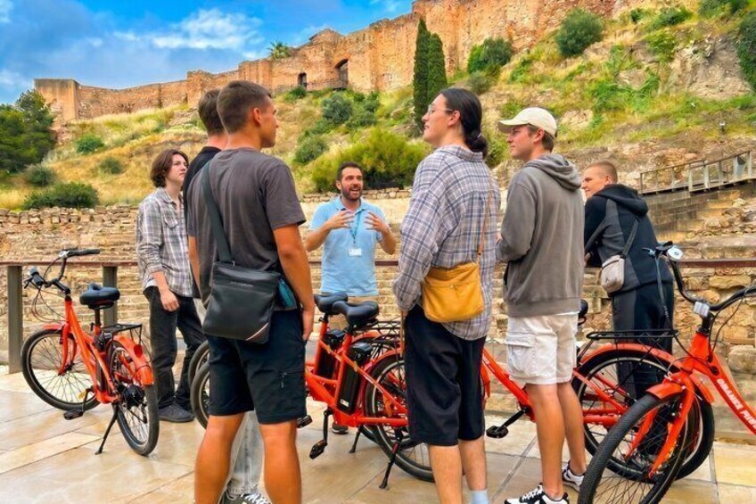 A group of people standing and talking near bicycles, with an ancient fort in the background. 2
