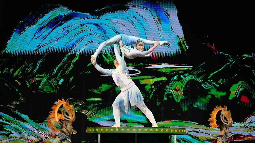 A performer in white costume executing a high-flying acrobatic maneuver with a partner, set against a backdrop of a large digital screen displaying abstract patterns. 1