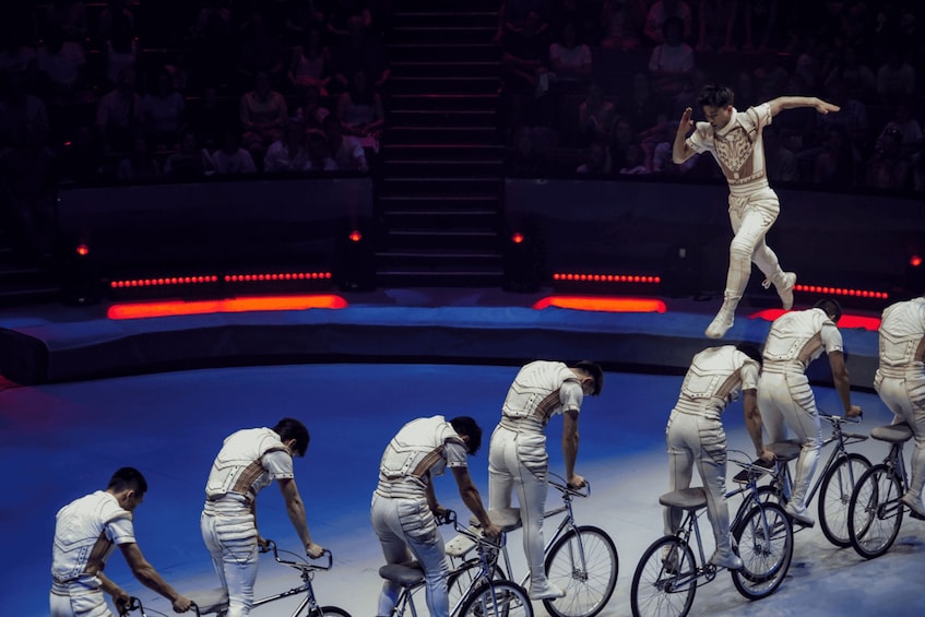 A group of performers in white costumes on bicycles, performing on a stage with an audience in the background. 5