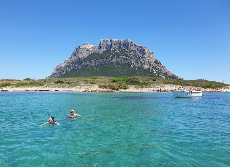 A clear blue sea with two people swimming, a rocky mountain in the background, and a boat on the water. 5