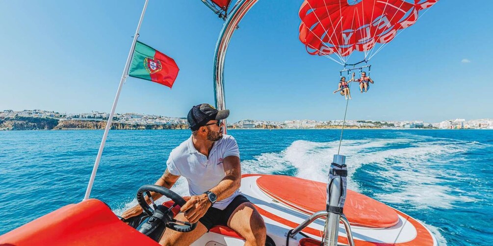 A man steering a red and white boat with a Portuguese flag, a parasail, and a coastal cityscape in the background. 1