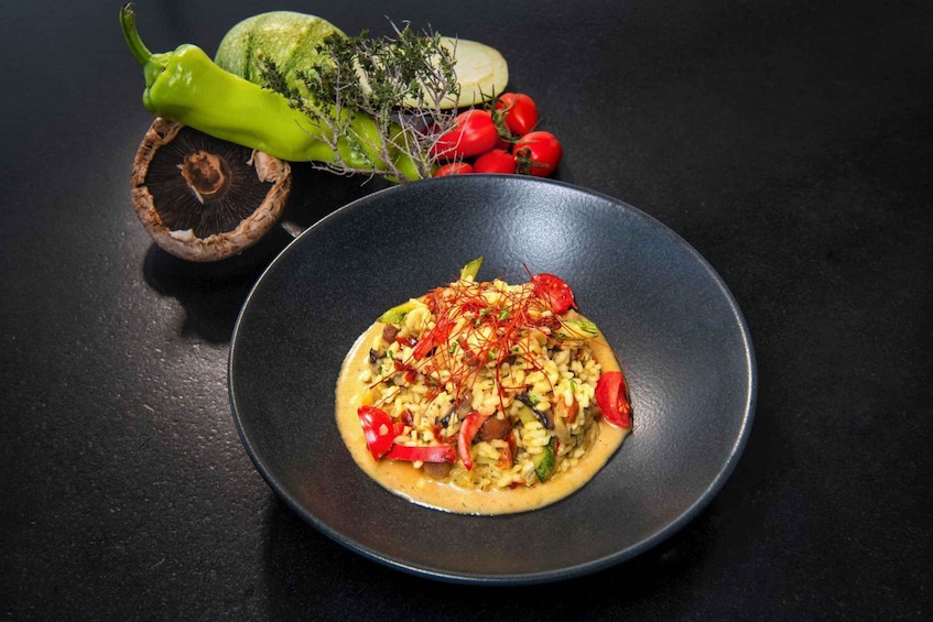 A dish with rice, vegetables, and red strands on a black plate, with a green pepper, cucumber, and tomatoes in the background. 5