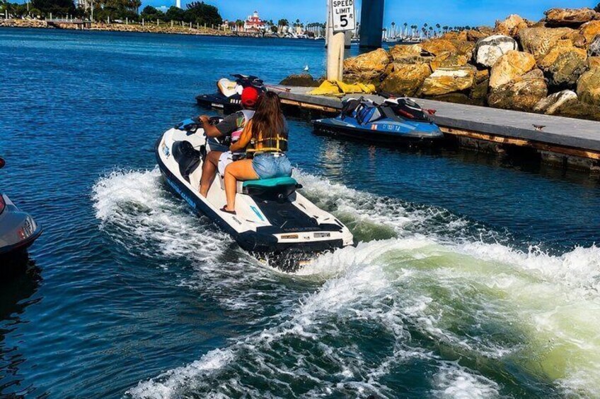 A jet ski with two riders on a body of water, a speed limit sign, and a rocky shoreline in the background. 4