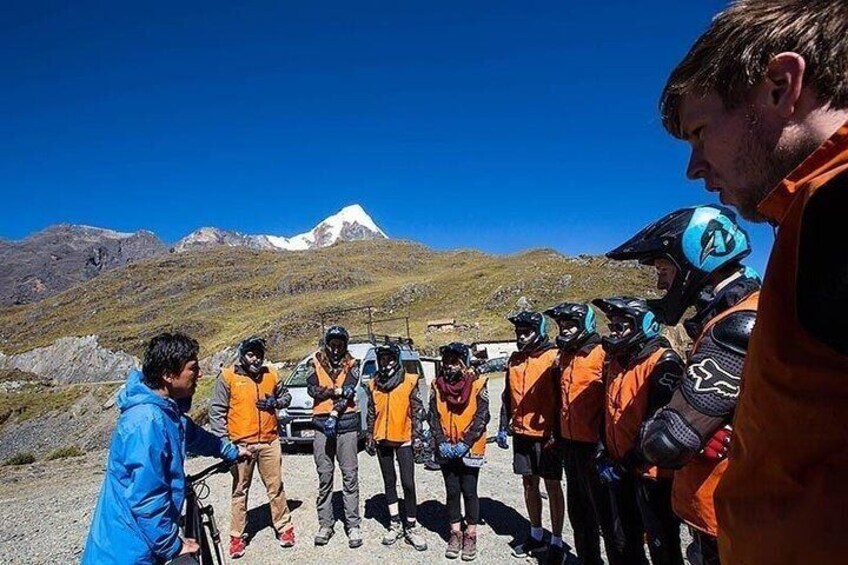 A group of people in orange vests standing on a mountain road with a clear blue sky and a snow-capped peak in the background. 3