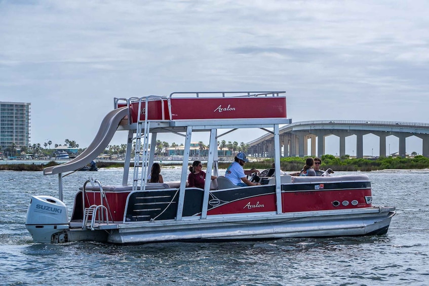 A red and white boat with a slide, named 'Avalon,' on a body of water with a bridge in the background. 1