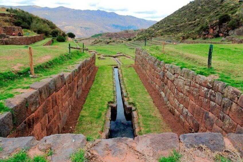 A stone-lined canal with a narrow waterway, flanked by grassy areas and stone walls, leading towards distant mountains. 1