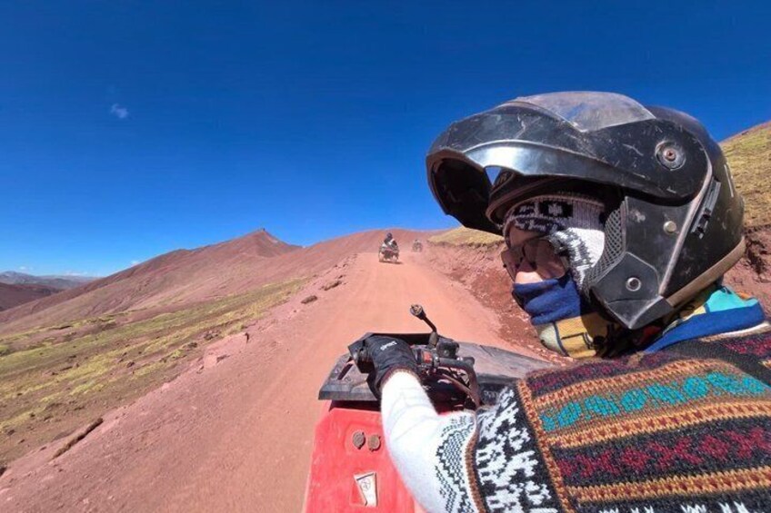 A motorcyclist on a red vehicle, wearing a helmet and gloves, on a dirt road with red hills in the background. 1